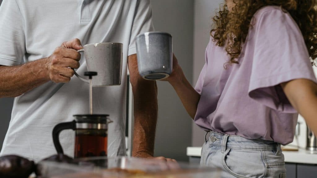 Two people standing in a kitchen holding mugs near a French press coffee maker on a counter.