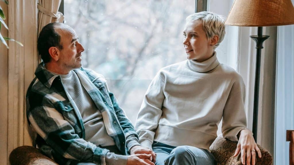 A man and woman sitting by a window, looking at each other while holding hands.