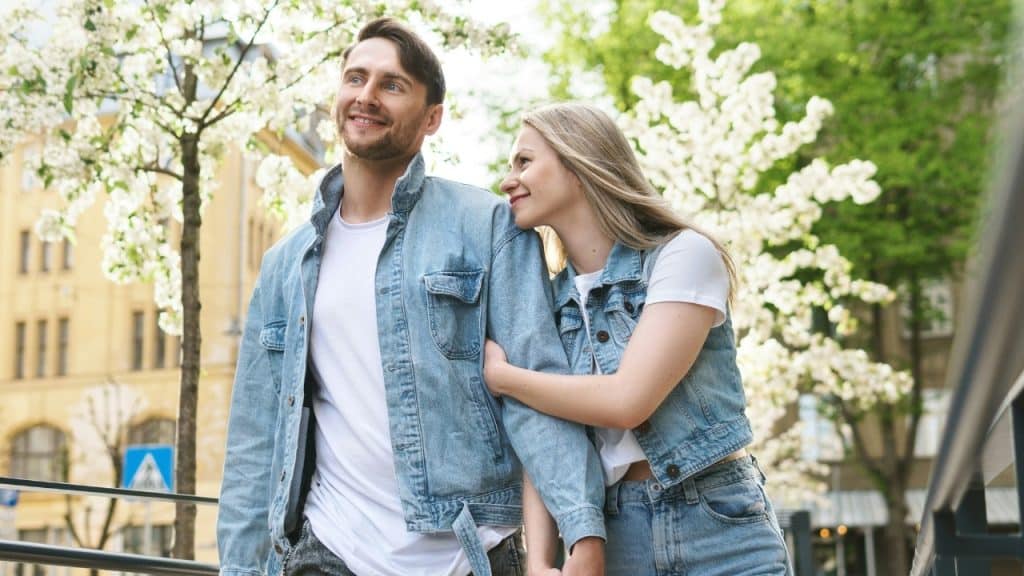 A man and woman wearing denim jackets walking outside past trees with white spring blossoms.