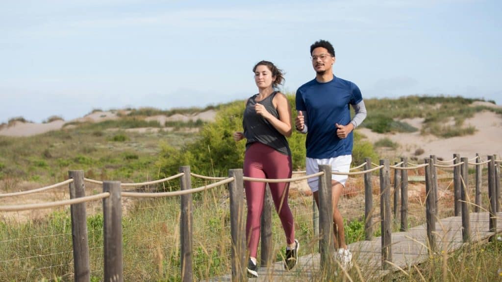 A man and woman jogging together across a wooden bridge in a grassy outdoor area.