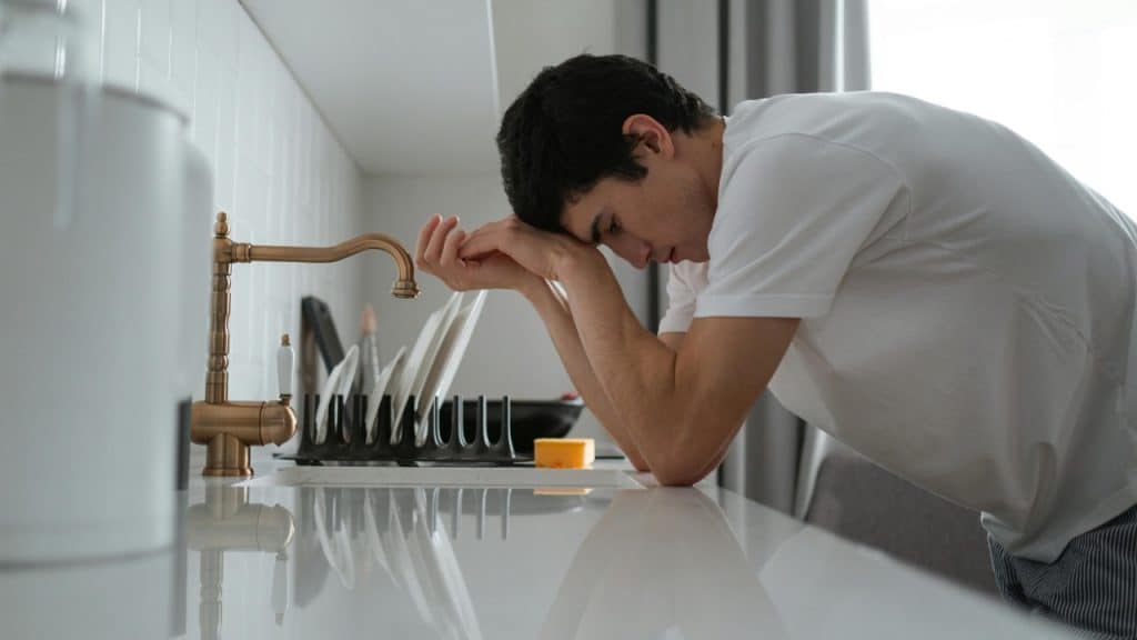 A man in a white t-shirt leans his head on his hands over a sink.
