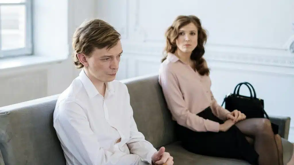 A man and woman sit far apart on a gray sofa in a white room.