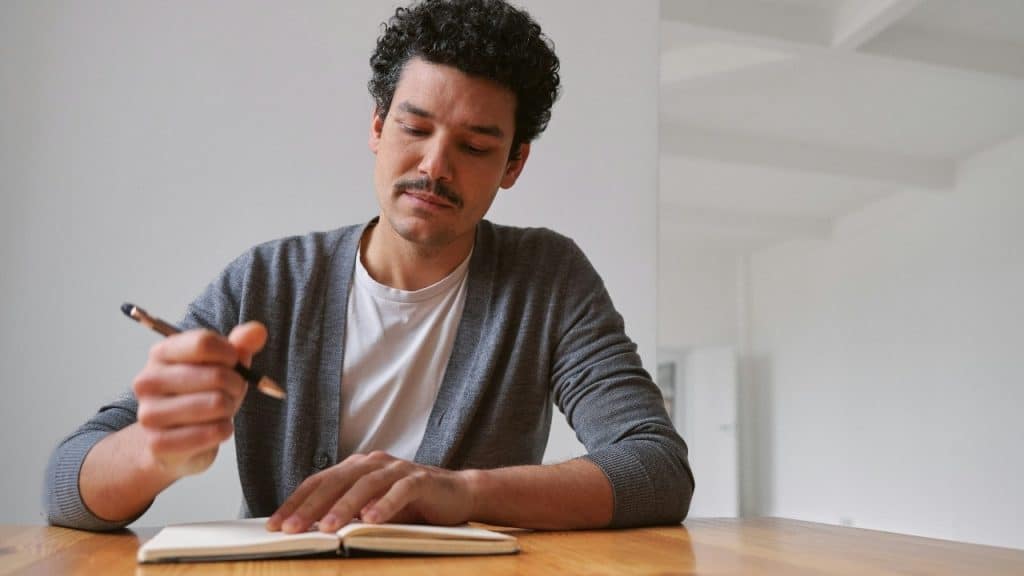 A man with curly hair and a mustache writes in a notebook at a table.