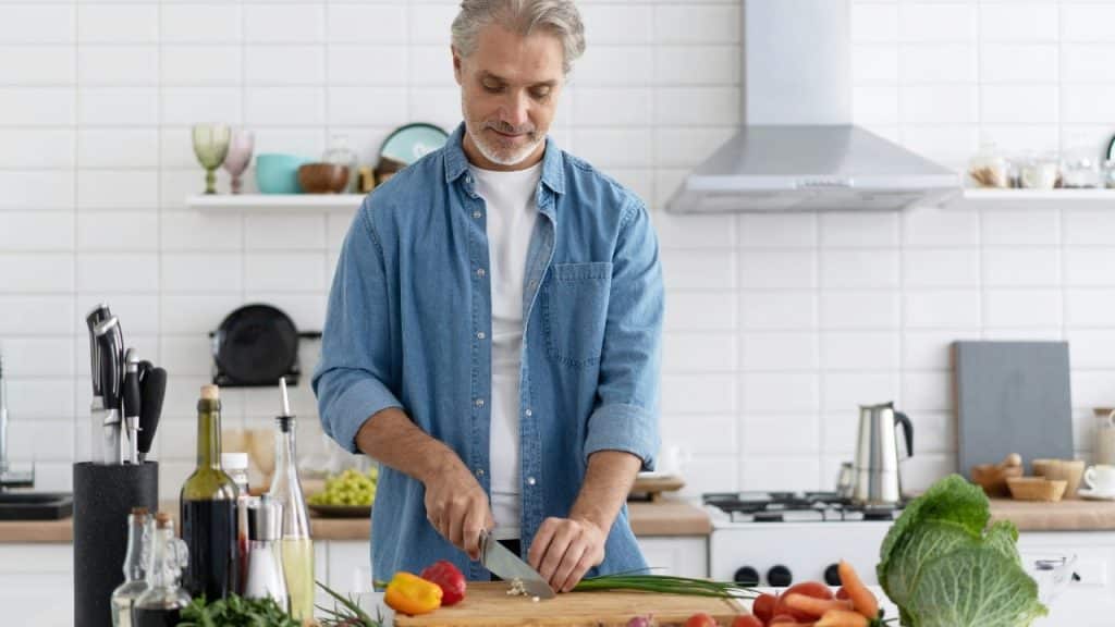 A man in a denim shirt slices green onions on a wooden cutting board.