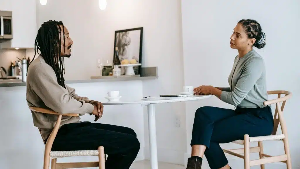 A man and woman sit at a round white table talking over coffee cups.