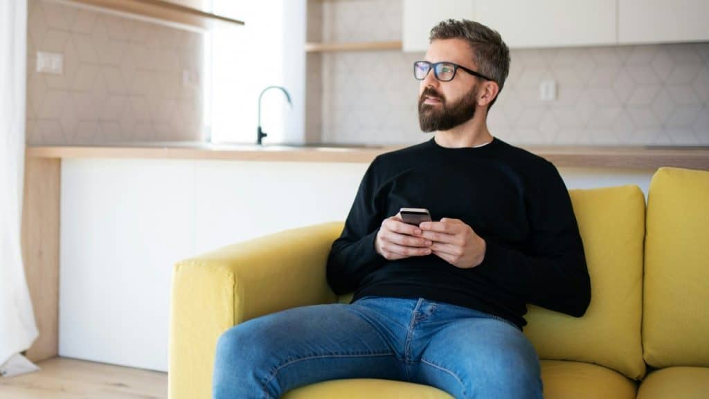 A man with a beard and glasses sits on a yellow sofa holding a smartphone.