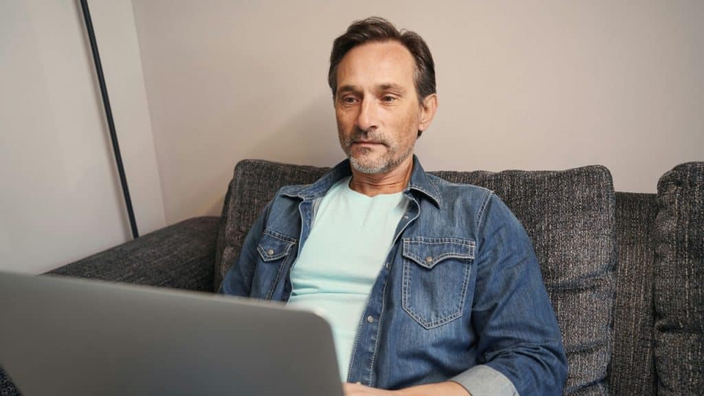 A man with a beard sits on a dark sofa looking at a laptop screen.