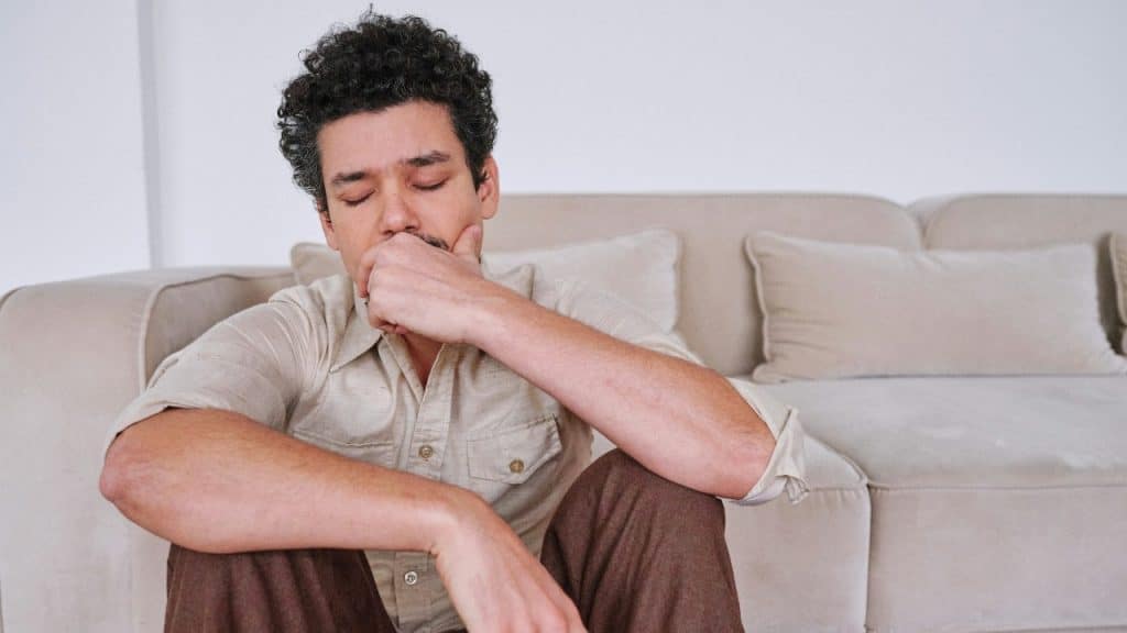 A man with curly hair sits on the floor, leaning his back against a beige sofa.