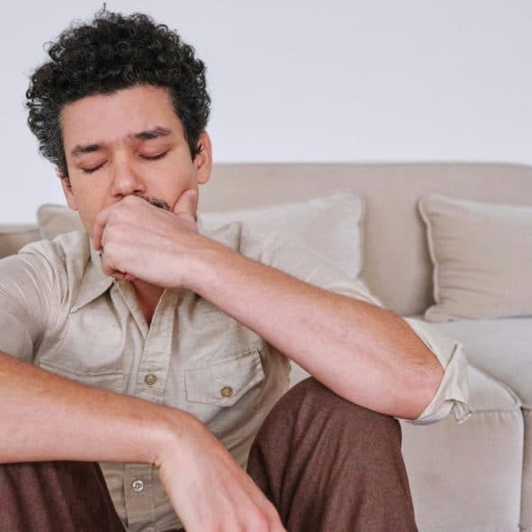 A man with curly hair sits on the floor, leaning his back against a beige sofa.