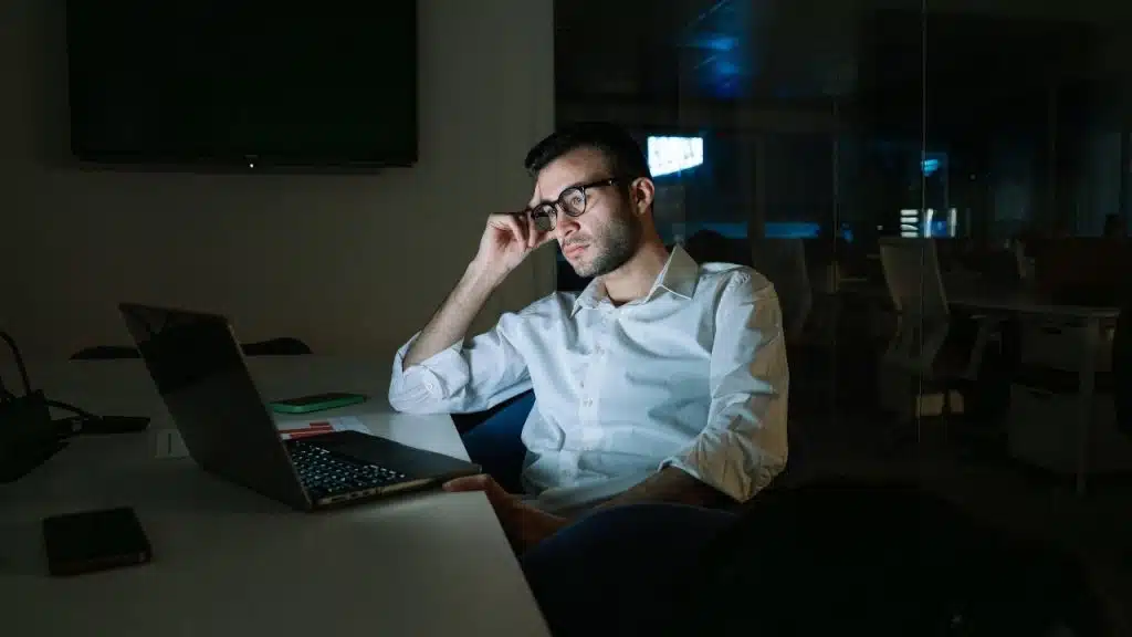 A man wearing glasses sits at a desk in a dark room working on a laptop computer.