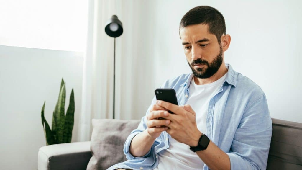 A bearded man in a light blue shirt sits on a grey sofa looking at a smartphone.