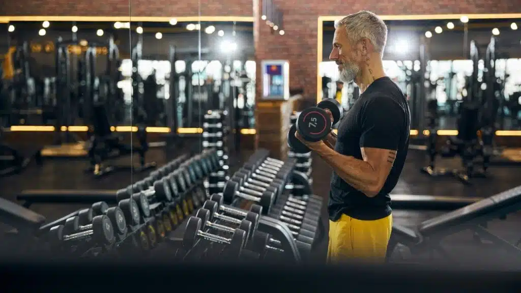 A bearded man in a gym lifts dumbbells while standing in front of a mirror and weight rack.