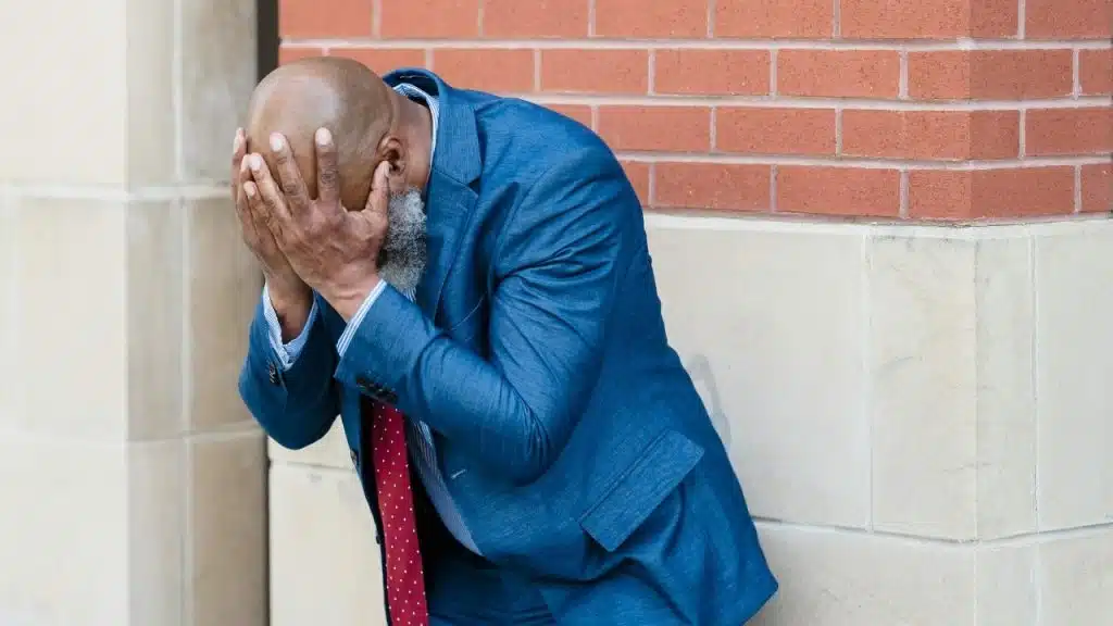 A man in a blue suit stands outside against a brick wall with his hands covering his face.