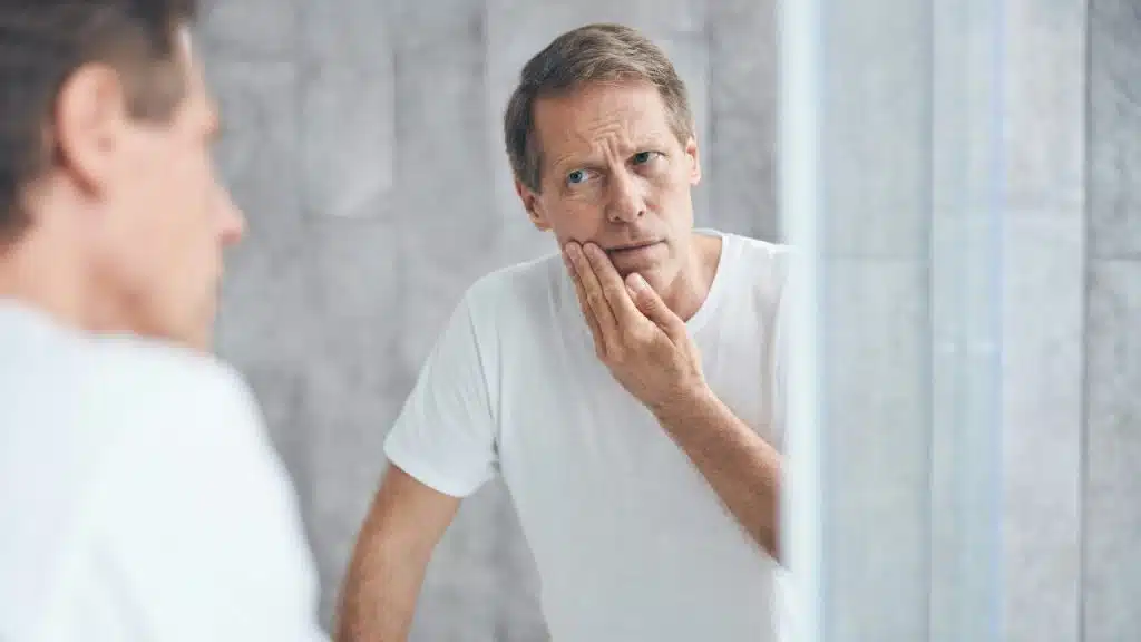 A man in a white t-shirt looks into a mirror while touching his hand to his face.