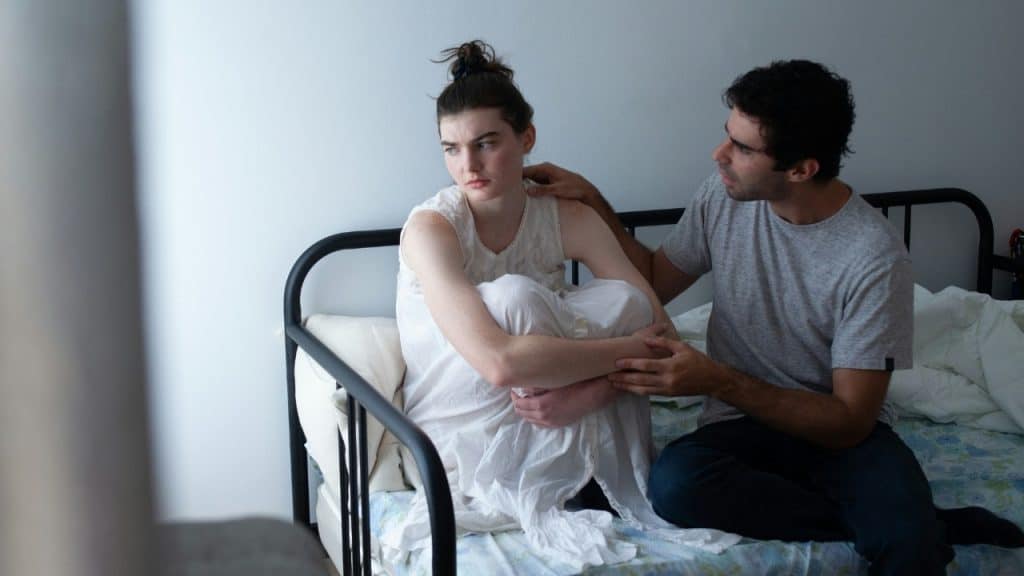 A man sits on a bed and places a hand on the shoulder of a woman looking away.