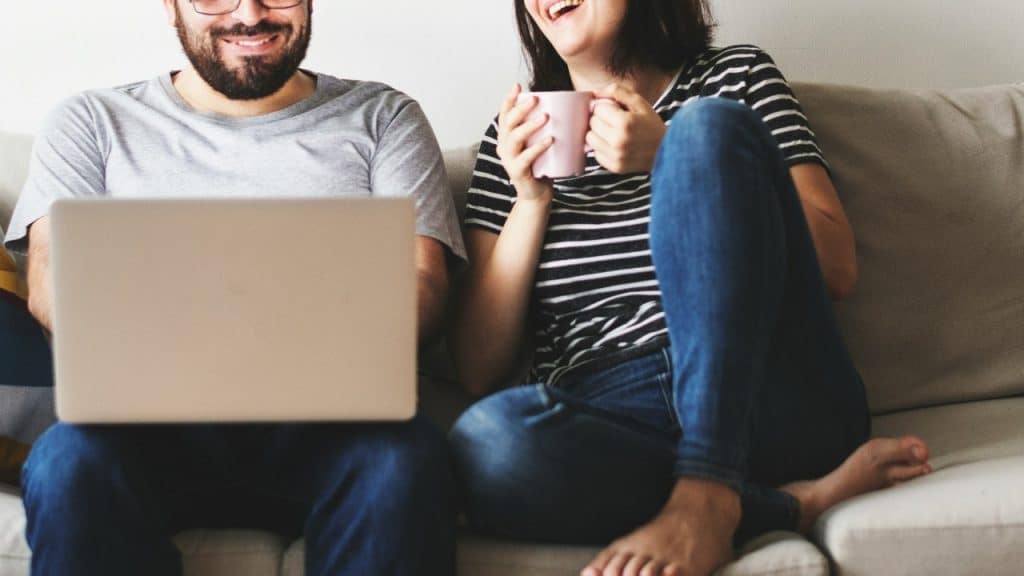 A man and woman sit on a couch together; one uses a laptop while the other laughs.