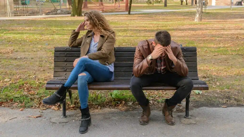 A woman and a man sit on a park bench facing away from each other with somber expressions.