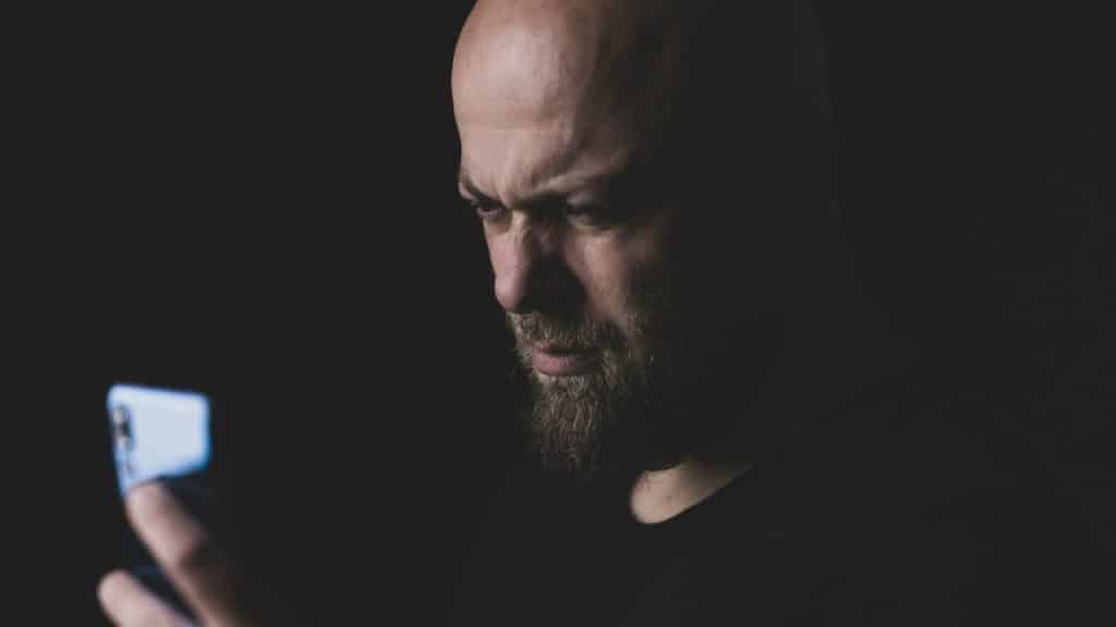 A bald man with a beard looks down at the glowing screen of a smartphone.