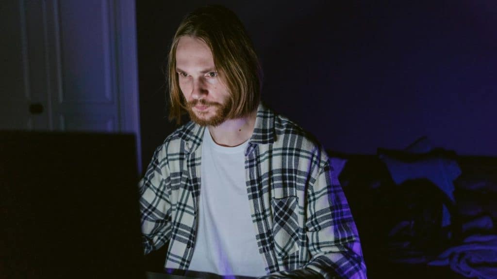 A man with long hair and a beard stares intently at a glowing laptop screen.