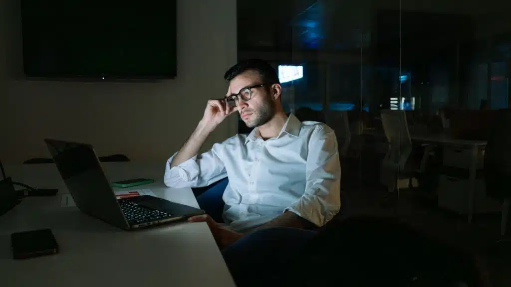 A man in glasses sits at a desk in a dark office, staring at a laptop.