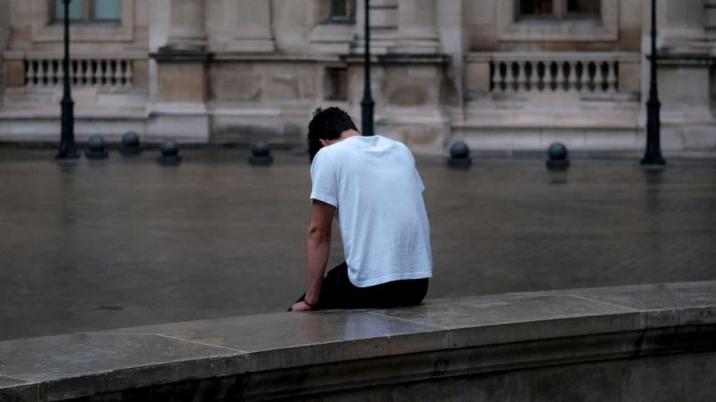 A person in a white t-shirt sits alone on a stone ledge facing a courtyard.