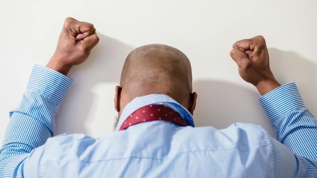 A man in a blue striped shirt leans his head and clenched fists against a wall.
