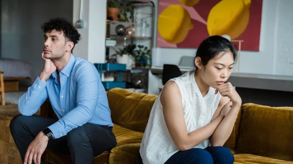 A man and woman sit on a yellow sofa, looking away from each other somberly.
