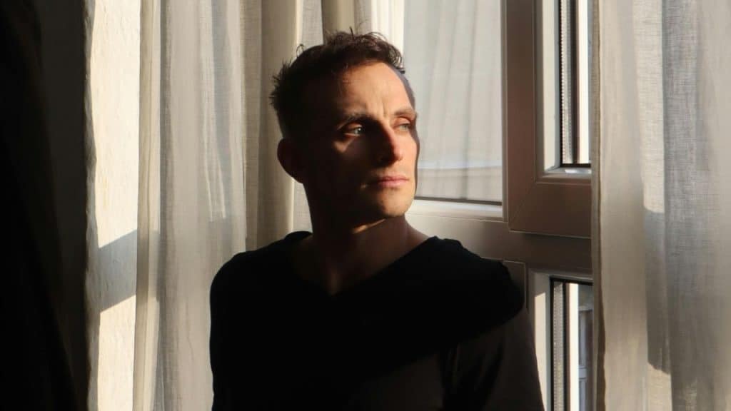 A man in a black shirt looks out of a window with sheer white curtains.