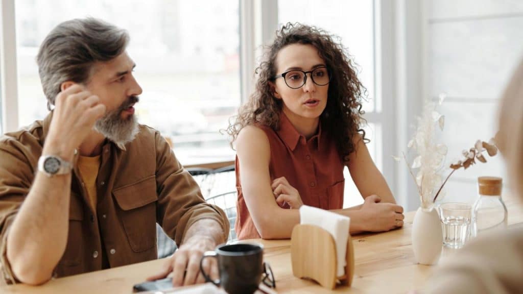 A man and woman sit at a wooden table during a conversation in a bright room.