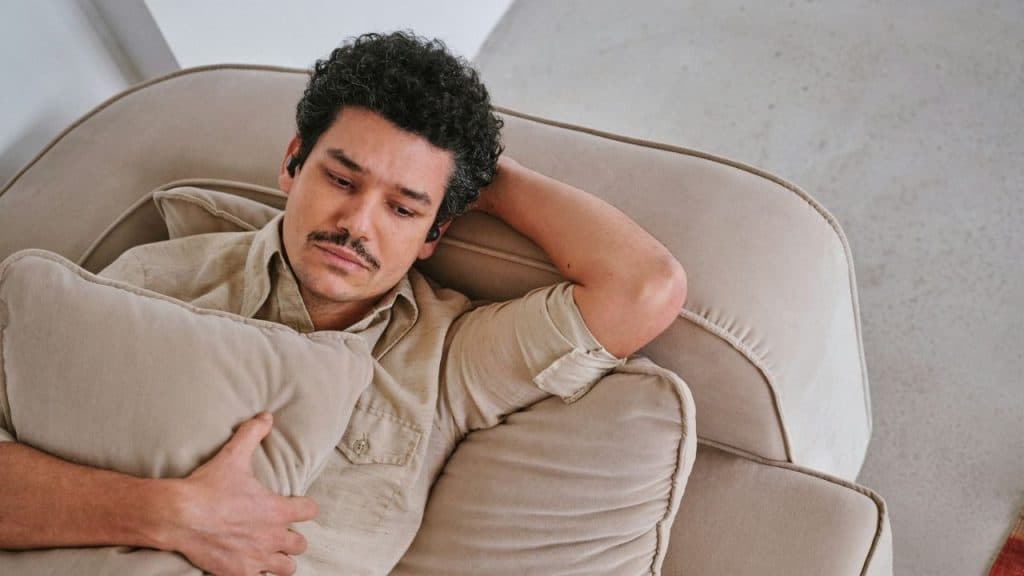 A man with curly hair and a mustache lies on a tan sofa holding a pillow.
