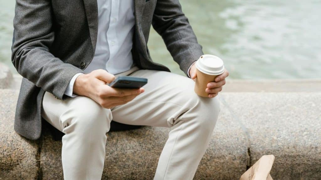 A person in a blazer sits on a stone ledge holding a phone and coffee.