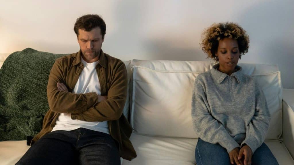 A man and woman sit on a white sofa, looking away with distant, somber expressions.
