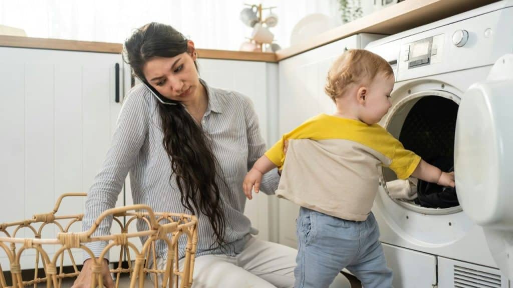 A woman on a phone holds a toddler reaching into an open washing machine drum.