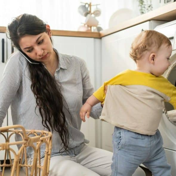 A woman on a phone holds a toddler reaching into an open washing machine drum.
