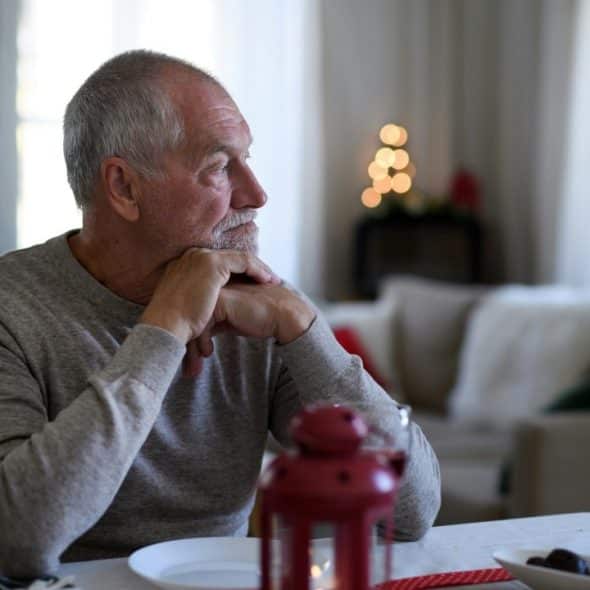 Senior man sitting at a table resting his chin on his hands, looking away thoughtfully.