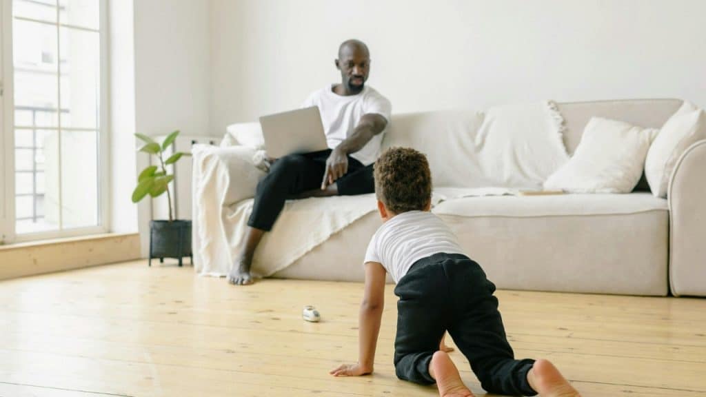 Man sitting on a sofa with a laptop looking at a child crawling on the floor.