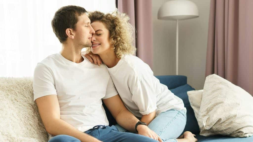 A woman leaning her head against a man's face while both sit on a sofa.