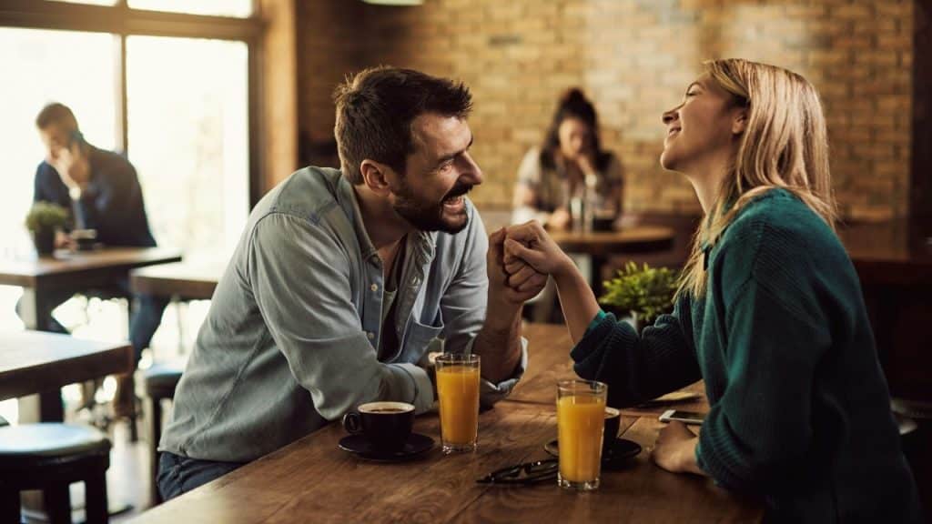 A man and woman sitting at a cafe table smiling and holding hands over drinks.