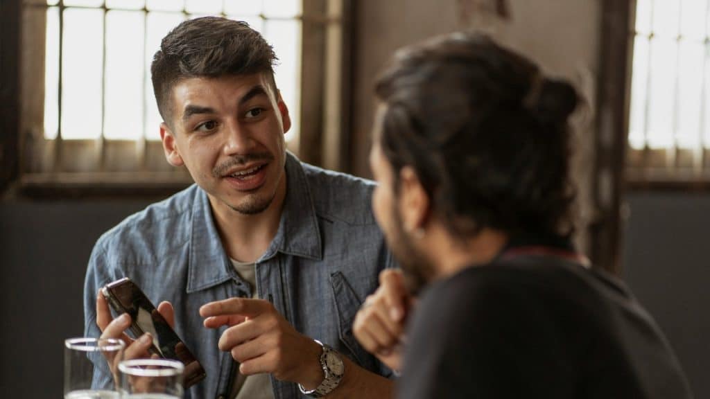 A man holding a smartphone and gesturing while talking to another man in a room.