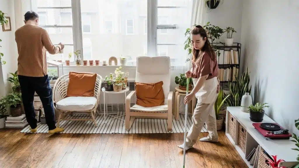 Man watering plants by a window while a woman mopping the floor in a room.