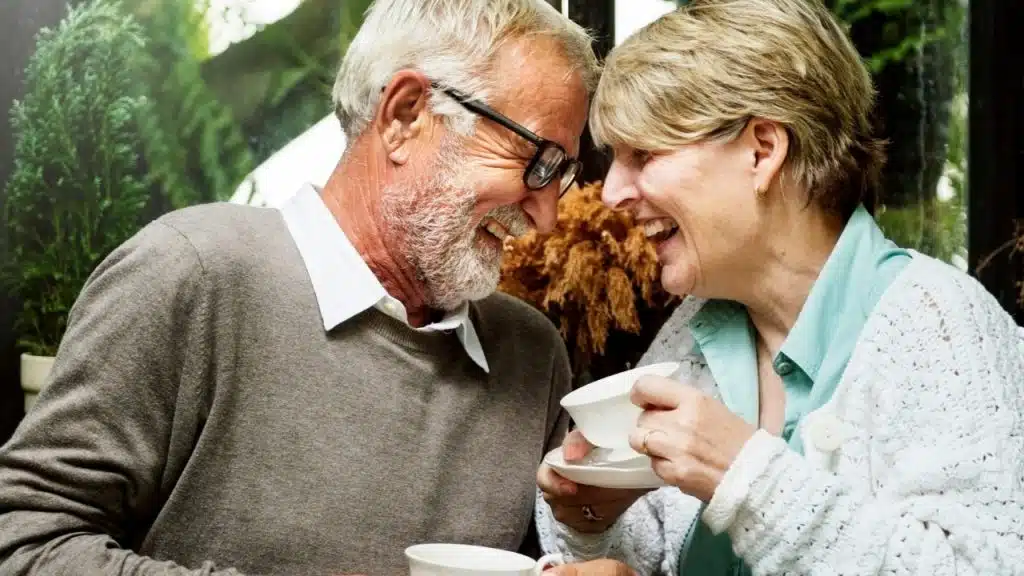 Senior man and woman laughing together while holding white teacups in a sunlit indoor setting.