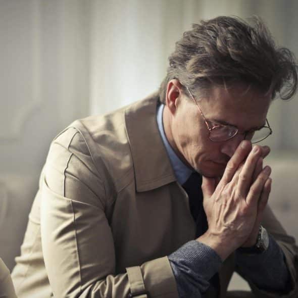 A man in a tan coat and glasses sits with his hands clasped near his face.