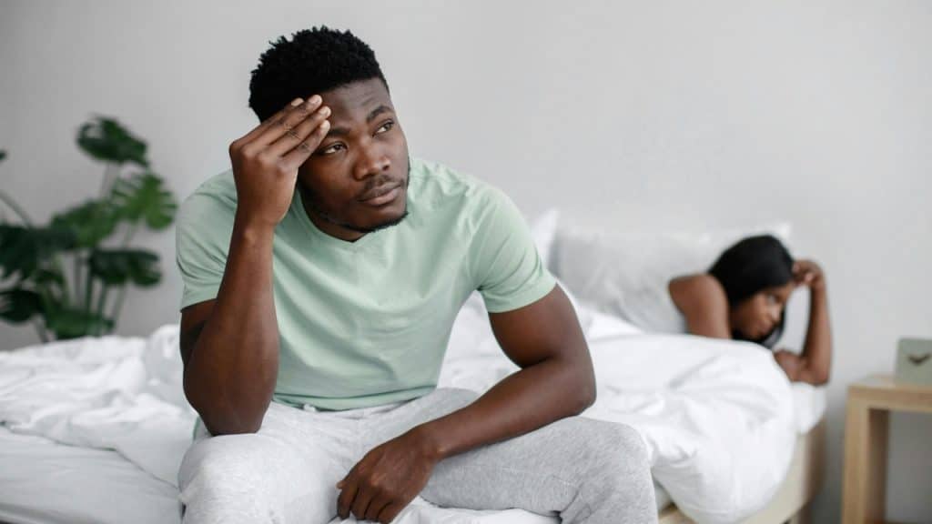 A man sits on a bed with his hand on his forehead near another person.