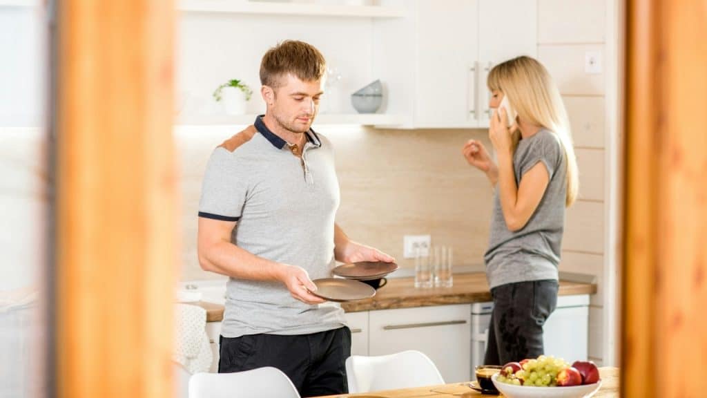 A man holds two plates in a kitchen while a woman talks on a cellphone.