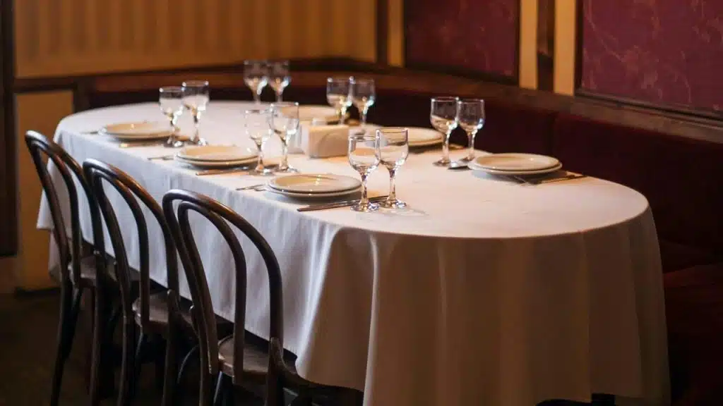 An empty long table with a white cloth is set with several plates and wine glasses.