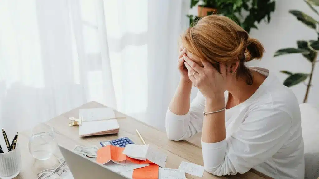 A woman sits at a desk with her face in her hands, surrounded by bills.