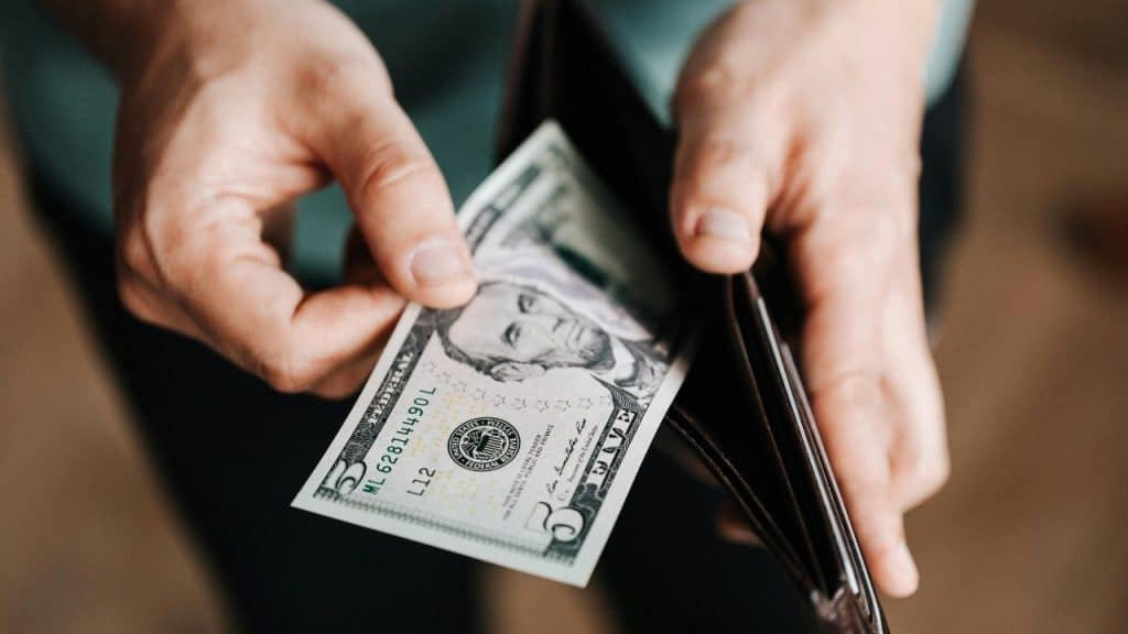 Close-up of hands pulling a five-dollar bill out of a brown leather wallet indoors.