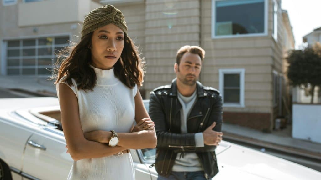 A woman and man stand with arms crossed near a white car on a street.