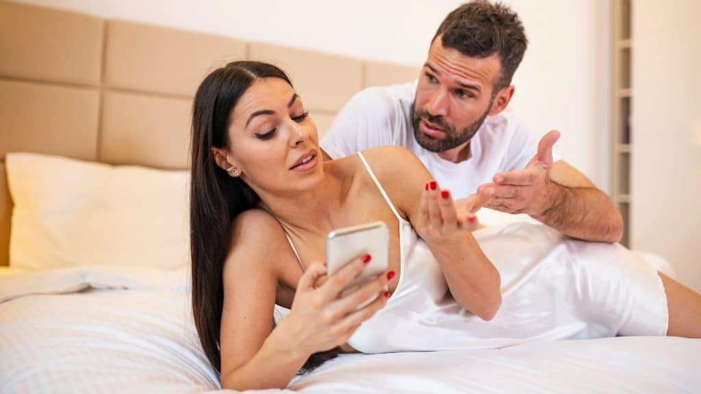 A woman lies on a bed looking at a phone while a man gestures toward her.