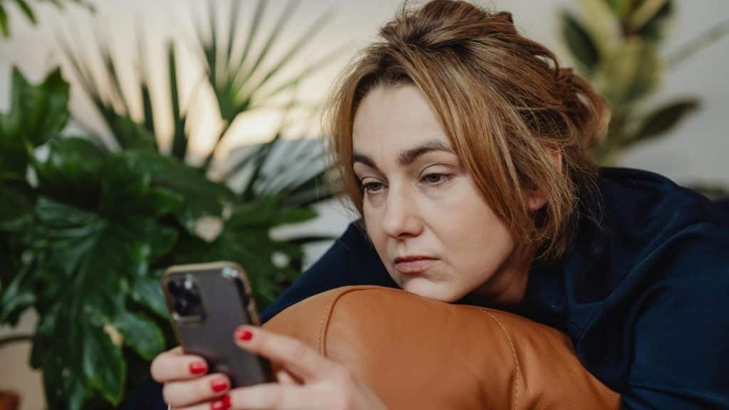 A woman with tied-back hair rests her chin on a cushion while looking at a smartphone.